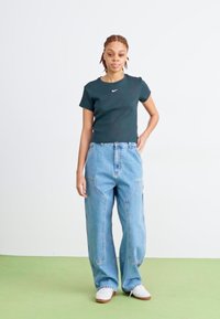 Juodas trumparankovis Nike marškinėliai derinami su šviesiai mėlynomis plačiomis džinsomis. Modelis dėvi baltus batus. Paprastas, švarus dizainas be matomų raštų.