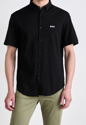 Man wearing a black short-sleeve button-up shirt with "BOSS" logo on chest and light olive pants, standing against a white background.
