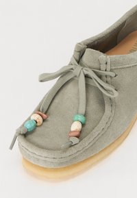 Chaussure en daim gris avec un bout rond, des lacets élastiques et des perles décoratives en turquoise et tons terre. Semelle en caoutchouc claire.