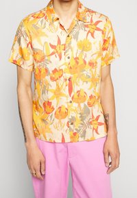 Camicia a maniche corte in un leggero tessuto beige, caratterizzata da un motivo floreale colorato con tonalità di rosso, giallo e verde, con chiusura a bottoni e vestibilità comoda.