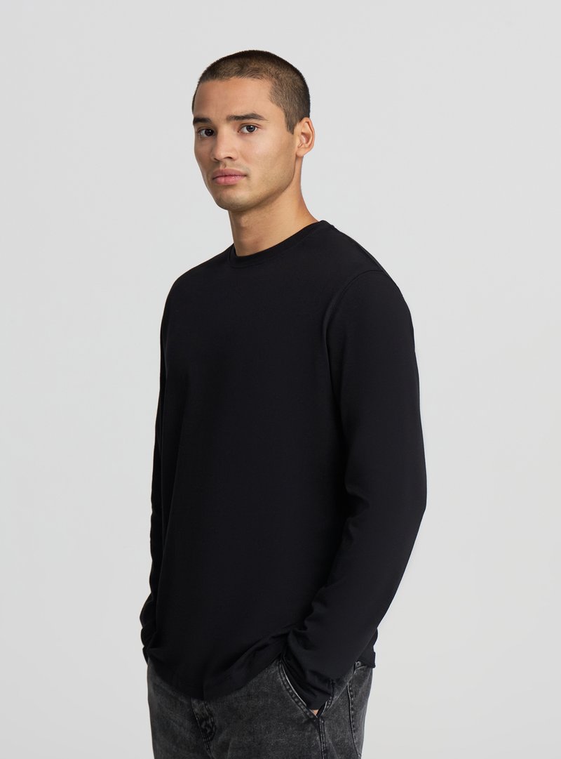 Giovane uomo con capelli corti che indossa una maglia a maniche lunghe nera semplice e jeans scuri, in piedi con le mani nelle tasche davanti a uno sfondo grigio.