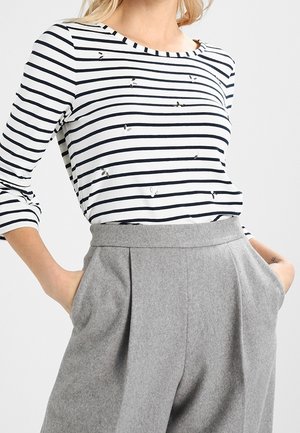 Kobieta ubrana w biało-czarną koszulkę z długim rękawem w paski z małymi motylkami oraz szare wysokowalne plisowane spodnie, z rękami w kieszeniach.