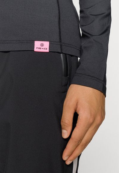 Camisa desportiva preta de mangas longas com um rótulo rosa. Apresenta costuras planas, um design justo e um bolso lateral com fecho zip parcialmente visível.