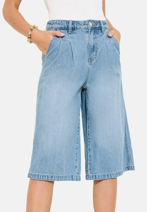 Lyseblå cowboyshorts i knælængde med forreste læg, sidelommer og knappelukning, båret af en person med hænderne i lommerne.