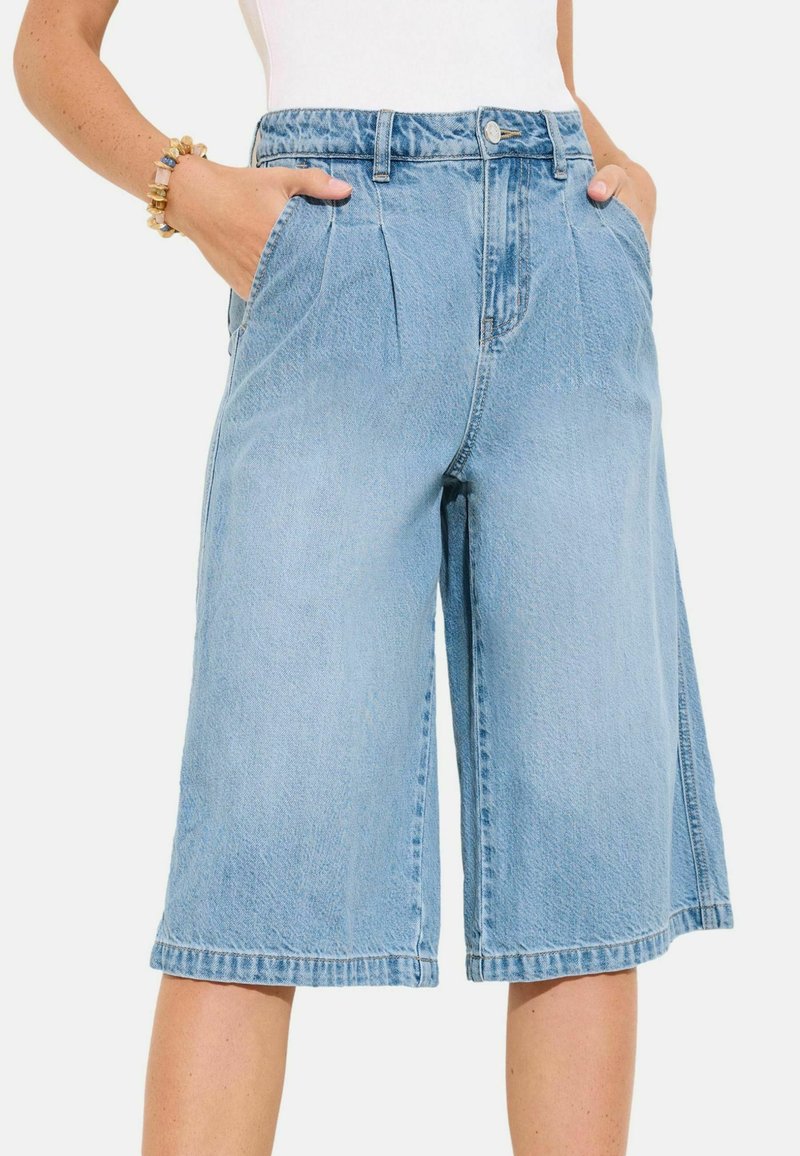 Hele sinised teksapõlvpüksid, millel on esipostitused, küljetaskud ja nööpkinnitus, kantud inimesel, kellel on käed taskutes.