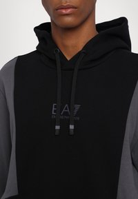 Svart och grå hoodie med en logotyp, dragsko med svarta spetsar och färgblockdesign på ärmarna. Mjuk tygtextur.