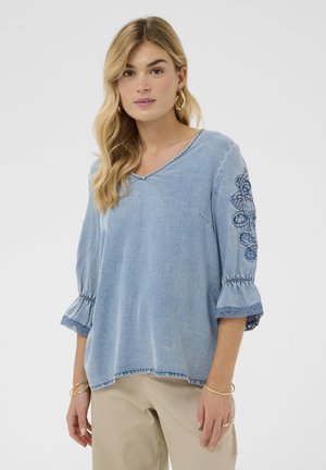 Žena s vlnitými blond vlasmi nosí svetlomodrú vyšívanú rifľovú blúzku s 3/4 rukávmi a béžové nohavice na jednofarebnom bielom pozadí.