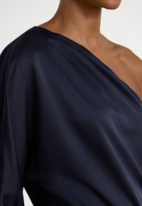 Ναυτικό μπλε σατέν τοπ με ασύμμετρο σχέδιο που αφήνει ακάλυπτο έναν ώμο, με λεία υφή και ύφασμα που πέφτει απαλά για κομψή κίνηση.