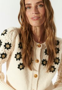 Cardigan lavorato a maglia avorio con ricamo floreale in nero e beige, con un motivo testurizzato sulle maniche e bottoni in legno.