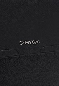 Portafoglio in pelle nera con texture e dettaglio del logo "Calvin Klein" in argento. Caratterizzato da un design minimalista e accenti lisci.