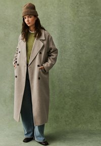 Grijze dubbelgeknoopte wollen jas met grote zwarte knopen, zijzakken, over een groene trui, gecombineerd met uitlopende lichtblauwe jeans en donkere schoenen.