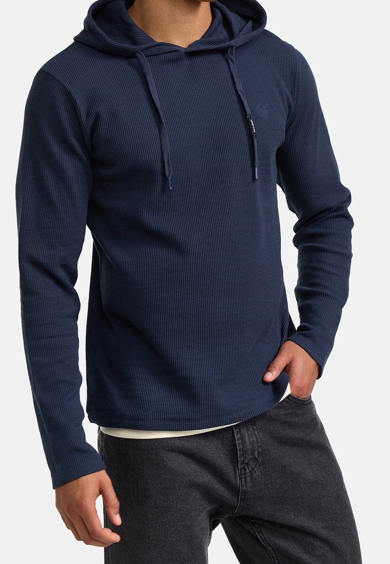 Sudadera marina con textura acanalada, que presenta un bolsillo frontal y cordones ajustables en la capucha. Combinada con jeans oscuros.