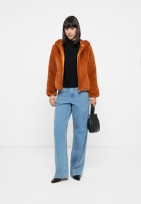 Synthetisch bont oranje jas met capuchon, rits aan de voorkant en elastische manchetten. Gecombineerd met lichtblauwe wijde jeans en een kleine zwarte tas.