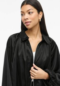 Černá saténová bomber bunda se zipem, řasené detaily na ramenech a balónovými rukávy; hladká textura a minimalistický design.