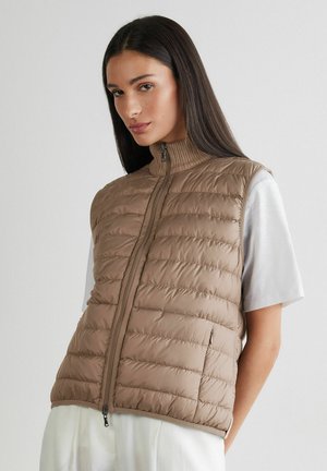 Junge Frau mit langem dunklem Haar, die eine beige gesteppte ärmellose Weste über einem weißen Kurzarmshirt trägt und vor einem einfarbigen Hintergrund steht.