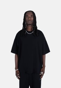 Black oversized t-shirt in soft fabric with short sleeves. Features a round neckline, minimal branding, and is paired with black trousers.