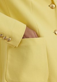 Χέρι στην τσέπη ενός κίτρινου σακακιού με υφή, με χρυσά διακοσμητικά κουμπιά στο μανίκι και στο μπροστινό μέρος.