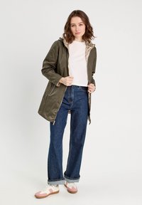 Veste à capuche vert olive avec une doublure métallique, des zips, des poches latérales, portée sur une chemise blanche et un jean évasé bleu, associée à des baskets roses.