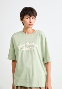 T-shirt oversized vert clair en coton doux, avec un texte en cursive blanc "New Balance Athletics" sur la poitrine. Manches courtes.