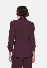 Donna con capelli castani corti, vestita con un blazer a maniche lunghe viola e pantaloni abbinati, vista di spalle su uno sfondo bianco.