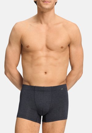 Hombre sin camiseta de pie con las manos detrás de la espalda, vistiendo calzoncillos bóxer ajustados de color gris oscuro sobre un fondo claro y liso.