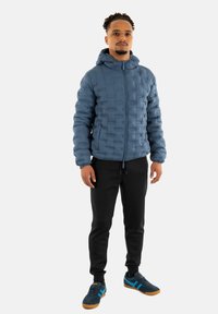 Giacca blu imbottita con zip frontale e cappuccio, caratterizzata da un motivo testurizzato. Pantaloni neri e scarpe scure con dettagli blu completano l'outfit.