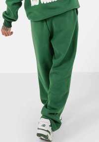 Groene, loszittende sweatpants met een gladde textuur, een elastische tailleband en enkelmanchetten, gecombineerd met witte sneakers met groene accenten.