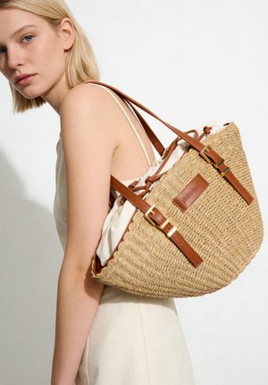 Femme blonde en robe légère portant un grand sac cabas en paille tressée avec des anses en cuir marron et un intérieur en tissu avec cordon de serrage.