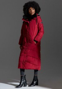 Red padded long coat with black faux fur trim, zippered front, and adjustable hood. Paired with black heeled ankle boots and dark jeans.