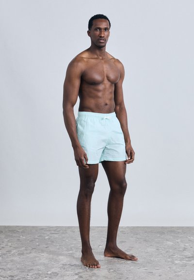 Αθλητικός άνδρας στέκεται ξυπόλυτος σε γκρι δάπεδο, φορώντας ανοιχτό μπλε μαγιό, μπροστά σε απλό ανοιχτό γκρι φόντο.