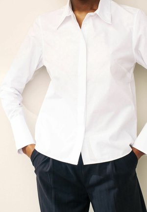 Persoon die een witte blouse met lange mouwen en zwarte broek draagt, met handen in de broekzakken, tegen een lichte achtergrond.