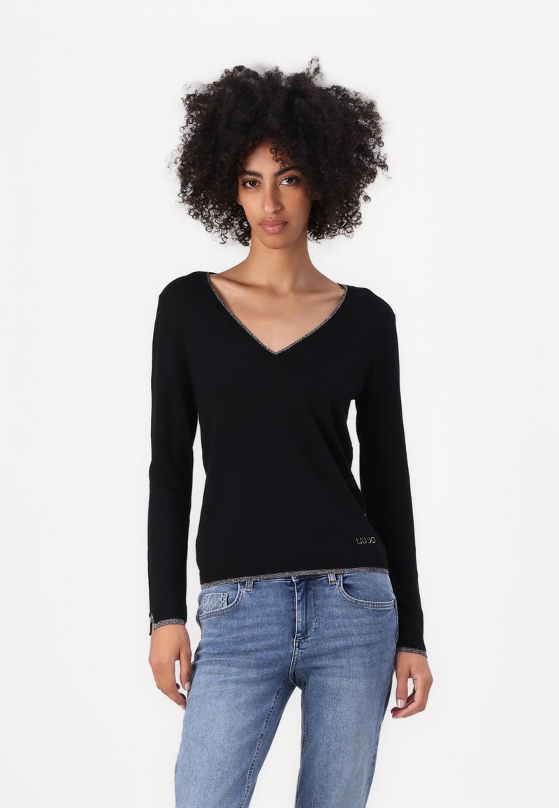Maglione nero a maniche lunghe con scollo a V e bordo argentato attorno al collo e all'orlo. Realizzato in un materiale morbido, abbinato a jeans blu.