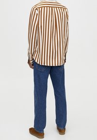 Personne debout portant une chemise à manches longues à rayures verticales marron et blanches, un jean bleu et des chaussures marron, dos tourné vers un fond uni.