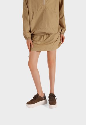 Donna che indossa una mini gonna color beige e un giubbotto abbinato con una grande tasca anteriore, abbinati a scarpe da ginnastica con zeppa marroni e suole bianche.