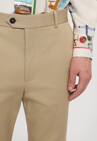 Beige Hosen mit schmalem Schnitt, aus strukturiertem Stoff. Mit einer Fronttasche mit sichtbaren Nähten und einem einzelnen schwarzen Knopfverschluss.
