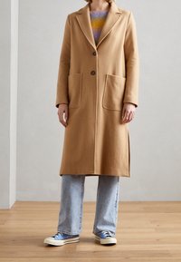 Kamelowy płaszcz wełniany z wyłamiem, dwoma dużymi kieszeniami z przodu i pojedynczym zapięciem na guzik, noszony na kolorowym swetrze i dżinsach.