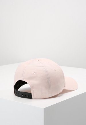 Casquette de baseball rose clair avec sangle noire réglable posée sur une surface d'angle blanche sur un fond clair uni.