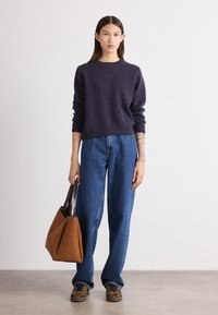 Marineblauer Strickpullover, hoher Kragen, lockere Passform. Helle blaue weit geschnittene Jeans, braune Wildleder-Tote, gemusterte Schuhe mit schwarzen und goldenen Akzenten.