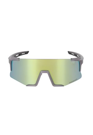 Nainen, jolla on pitkät laineikkaat hiukset, pukeutuneena vaaleansiniseen lyhythihainen paitaan ja heijastaviin ylisuurikokoisiin urheilulasien aurinkolaseihin, katsoo suoraan valkoisella taustalla.