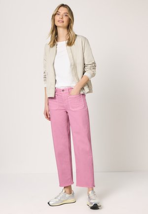 Junge Frau trägt eine beige Bomberjacke, ein weißes Hemd, pinke Jeans mit weitem Bein und silberne Sneakers, steht mit einer Hand in der Tasche.