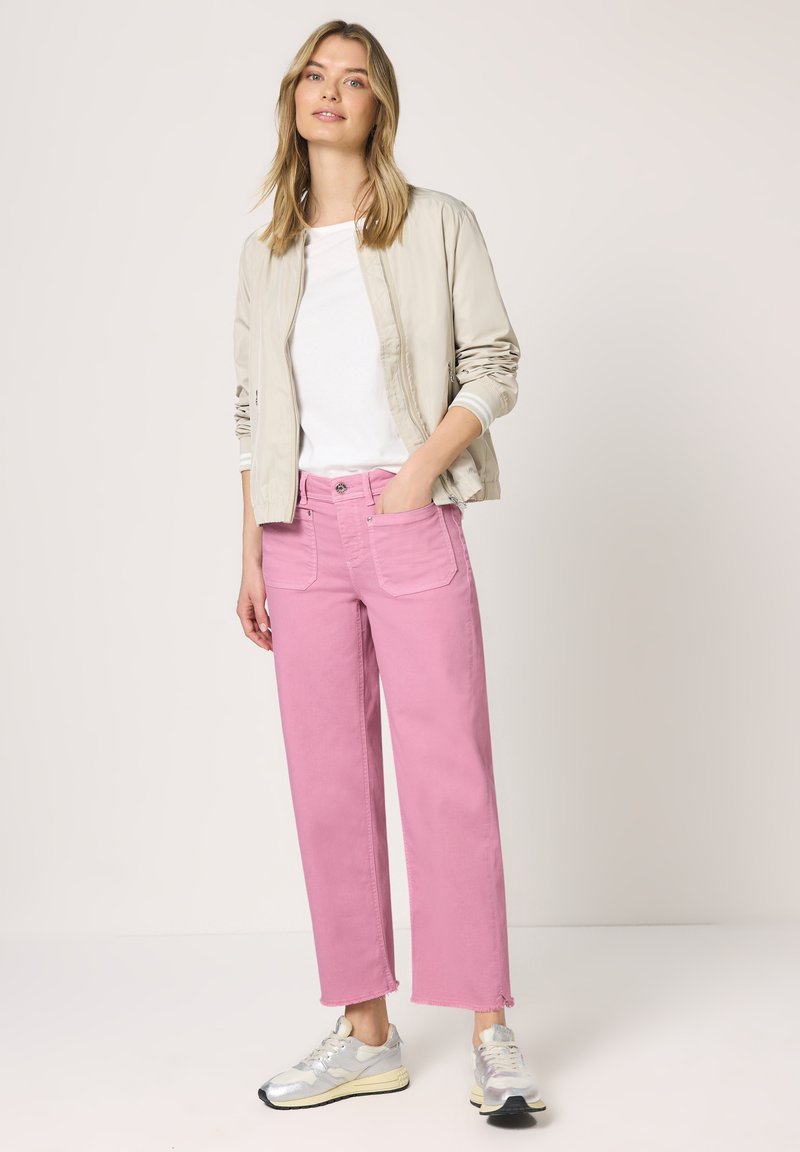 Jeune femme portant une veste bomber beige, une chemise blanche, un jean rose à jambes larges et des baskets argentées, debout avec une main dans la poche.