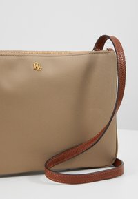 Borsa a tracolla in nylon khaki con una strap in pelle marrone. Presenta un logo dorato impresso e una chiusura con zip sulla parte superiore.