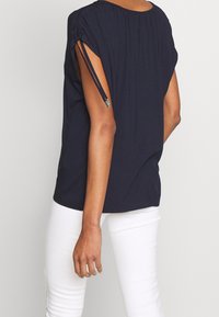 Námořnická modrá halenka s krátkým rukávem a nařasením na ramenech, s nastavitelnými šňůrkami, nošena s bílými kalhotami. Měkká, lehká látka.