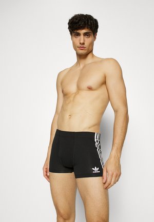 Shorts de bain noirs en tissu extensible, avec un design à trois rayures blanches sur les côtés et un petit logo à l'avant.