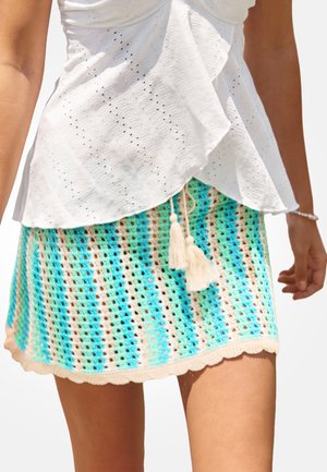 Mujer con una blusa blanca de ojales y una minifalda de crochet a rayas azul, verde y beige con flecos paseando al aire libre.