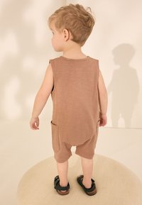 Brun ærmeløs romper med to sidelommer, blød tekstur og afslappet pasform. Kombineret med sorte sandaler, stående på en lys cirkulær overflade.