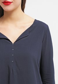 Blusa blu navy con scollo a V, maniche lunghe e bottoni decorativi argentati lungo la parte anteriore. Tessuto liscio con una consistenza morbida.