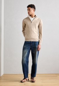 Beige, cipzáros nyakú kötött pulóver fehér galléros ing felett, világos kék farmernadrággal és barna bőrcipőkkel, kék fűzőkkel párosítva.