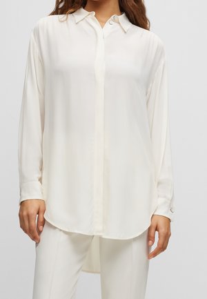 Vrouw draagt een oversized wit overhemd met verborgen knopen en witte broek, staand tegen een effen achtergrond.