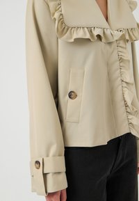 Blouse beige à manches longues avec col et devant à volants, manchette boutonnée et détail de bouton près de la taille, portée avec un pantalon noir.